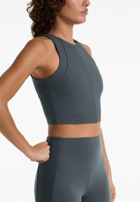 Femme portant un crop top sans manches à texture gris foncé et un legging taille haute assorti, se tenant avec une main sur la hanche.