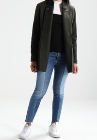 Tmavě zelený kabát s přiléhavým střihem, s detaily knoflíků a bočními kapsami, převlečený přes černý svetr a bílý rolák, v kombinaci s modrými džíny a bílými teniskami.