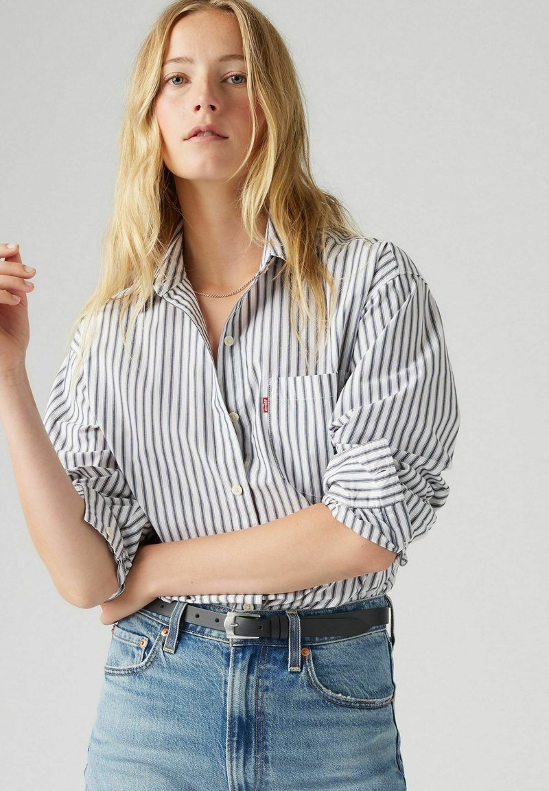 Pasiasta koszula z długim rękawem w kolorze białym i granatowym, z kieszonką na piersi i podwiniętymi rękawami, w połączeniu z jasnoniebieskimi dżinsami i czarnym paskiem.