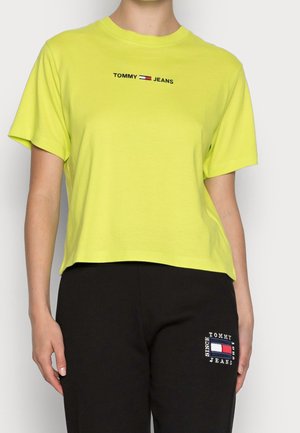 Osoba nosząca jasnoróżową krótką koszulkę z logo "TOMMY JEANS" i czarne spodnie z naszywką Tommy Jeans na udzie.