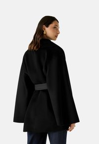 Cappotto nero con ampie maniche, tessuto strutturato, vita con cintura e colletto classico. Modello mostrato di spalle, indossa pantaloni di denim.