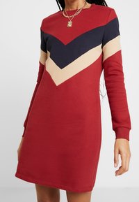 Robe-sweat rouge à manches longues, présentant un motif en chevron bleu marine, crème et bleu foncé sur la poitrine. Texture douce et encolure arrondie.