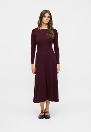 Νεαρή γυναίκα που στέκεται, φορώντας ένα μακρυμάνικο, ριμπ ύφασμα, μίντι φόρεμα σε σκούρο μοβ χρώμα και μαύρα μπαλαρίνα παπούτσια με δεσίματα στον αστράγαλο, σε απλό λευκό φόντο.