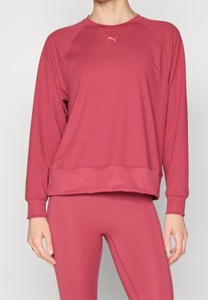 Kobieta ubrana w różowy długi rękaw bluzy Puma i dopasowane legginsy, stojąca na tle jednolitego, jasnego tła.