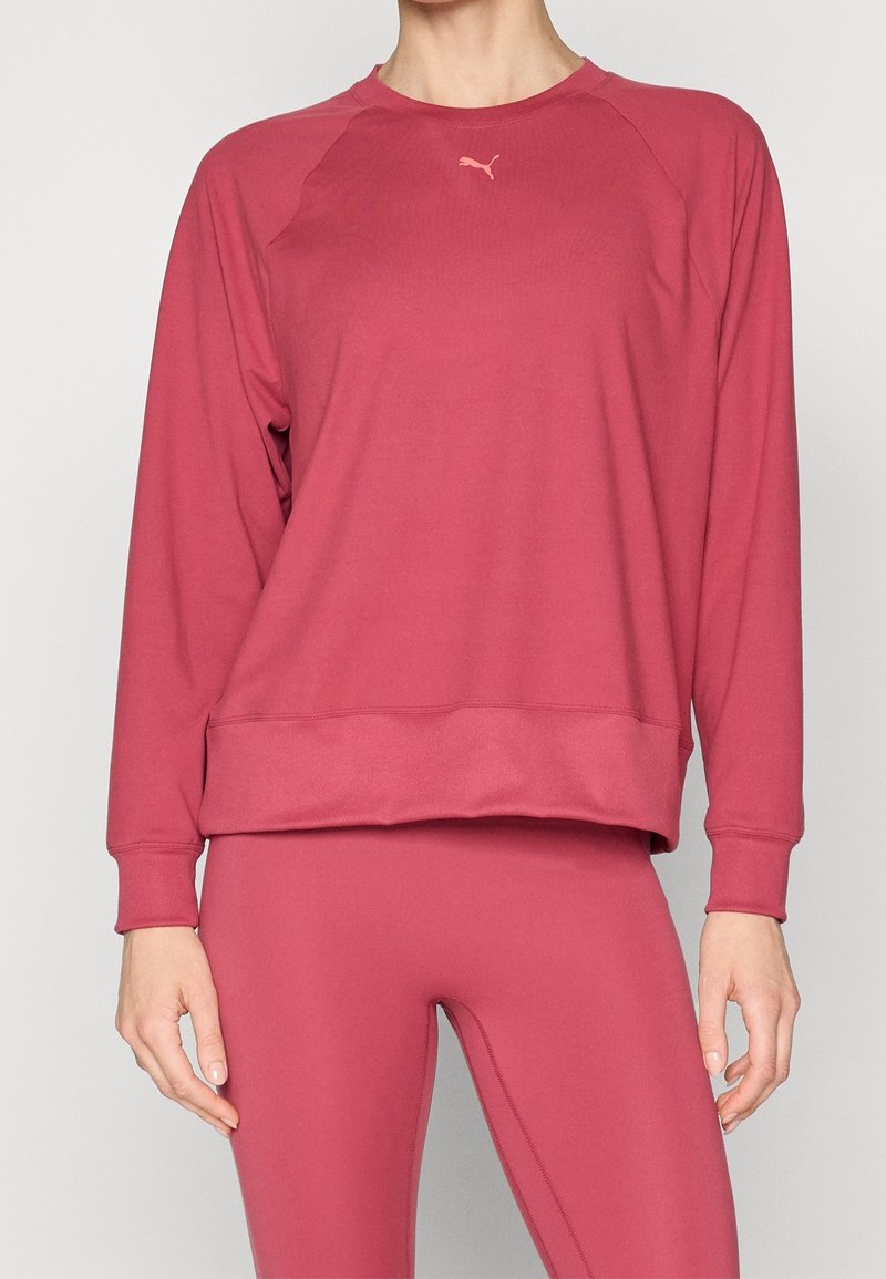 Femme portant un sweat-shirt Puma rose à manches longues et des leggings assortis, debout devant un fond clair uni.