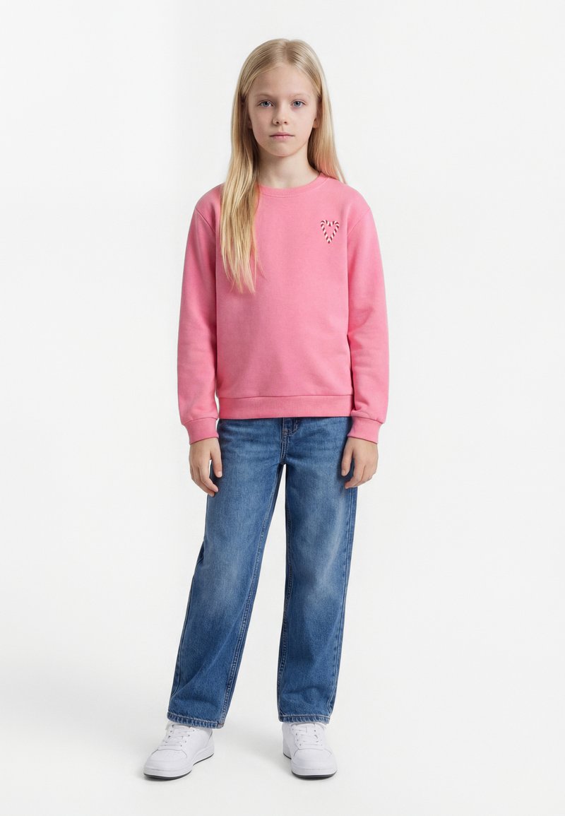 Hosszú szőke hajjal rendelkező lány áll, rózsaszín pulóvert visel szívmotívummal, kék farmert és fehér sportcipőt fehér háttér előtt.