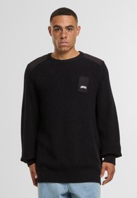 Maglione nero lavorato a maglia con polsini e orlo a coste, caratterizzato da un tessuto testurizzato e un patch quadrato con il logo "UNFAIR" sul petto.