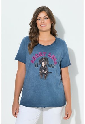 Kobieta w niebieskiej koszulce z grafiką jamnika i napisem "KRÓTKIE NOGI, BRAK OGRANICZEŃ", ubrana w białe spodnie, uśmiechająca się na białym tle.