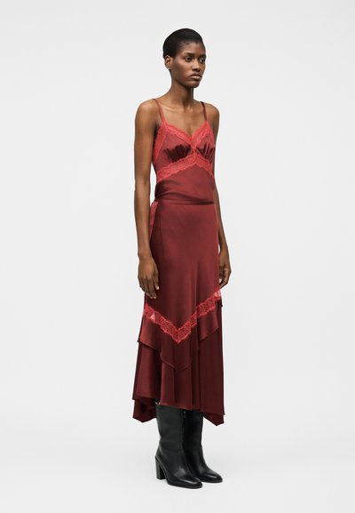 Vrouw staand, gekleed in een mouwloze bordeauxrode satijnen jurk met rode kanten rand, gecombineerd met zwarte laarzen met hak tot over de knie, op een witte achtergrond.