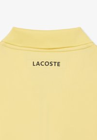 Polo giallo con colletto a costine, dotato di un'etichetta rettangolare sul retro con la scritta "LACOSTE" in testo nero. Texture liscia.