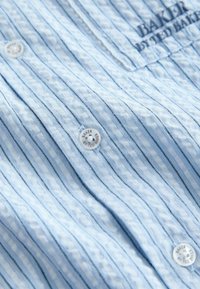 Chemise bleu clair texturée avec fines rayures verticales noires, boutons blancs de marque portant l'inscription "Baker by Ted Baker" et logo partiellement brodé sur la poitrine.