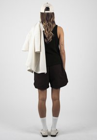 Femme aux longs cheveux ondulés portant un débardeur noir, un short noir, des chaussettes blanches, des chaussures et une casquette blanche, tenant une chemise rayée sur une épaule.