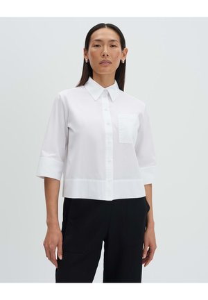 Frau mit glattem dunkelbraunem Haar trägt ein weißes Hemd mit Dreiviertelärmeln und schwarze Hose, steht vor einem schlichten hellen Hintergrund.