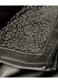 Schwarzes Lederkleidungsstück mit einem flauschigen, strukturierten Wollkragen. Der Kragen ist von glattem Leder mit kontrastierenden Nähten und einem Reißverschluss umrandet.