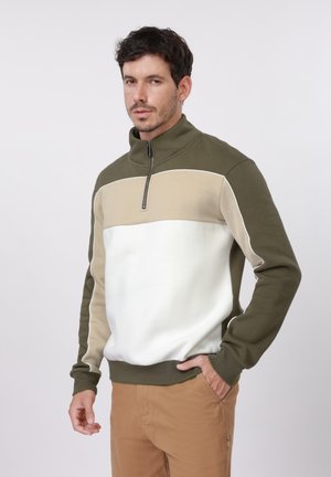 Muž nosiaci olivovo zelený, béžový a biely pulóver so štvrťzipsom, so založenou rukou v vrecku, stojaci pred jednoduchým sivým pozadím.