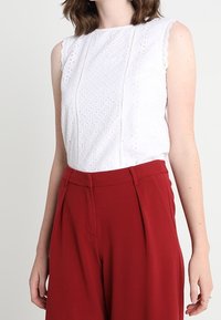Bílý bezramínkový top s vyšívanými detaily, s vroubkovanými okraji; spárován s vysokým pasem a širokými červenými kalhotami s přehyby v přední části.