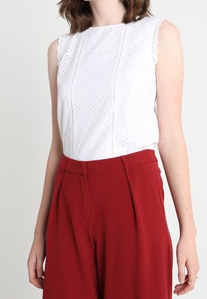 Top bianco senza maniche con ricamo ad occhio e bordi sagomati; abbinato a pantaloni rossi a vita alta e gamba larga, con pieghe frontali.