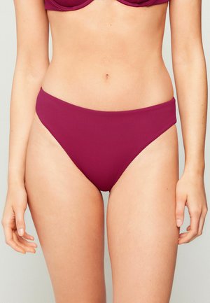 Kobieta w prążkowanych fioletowych majtkach od bikini i dopasowanym topie, widoczna od talii do górnej części ud na neutralnym tle.