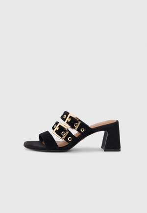 Black suede heeled mules featuring three wide straps with gold buckle accents and eyelets, round open toe, and chunky block heel.