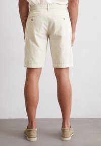 Light beige shorts made of textured fabric, featuring a straight cut and two back pockets with gold-tone button accents.