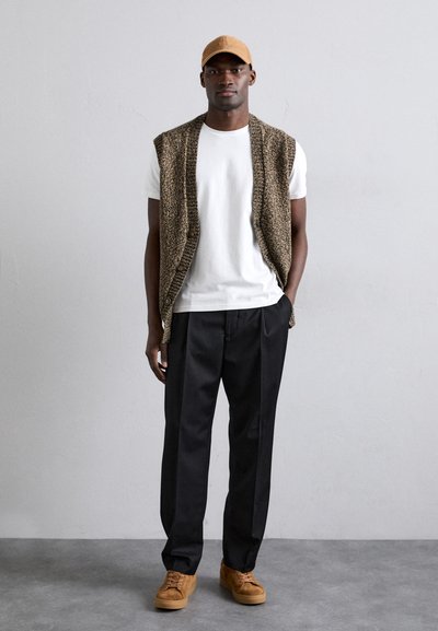 Gilet marrone sopra una maglietta bianca, abbinato a pantaloni neri e sneakers in suede color cammello. Design semplice con dettagli in pulsanti, texture minimale.