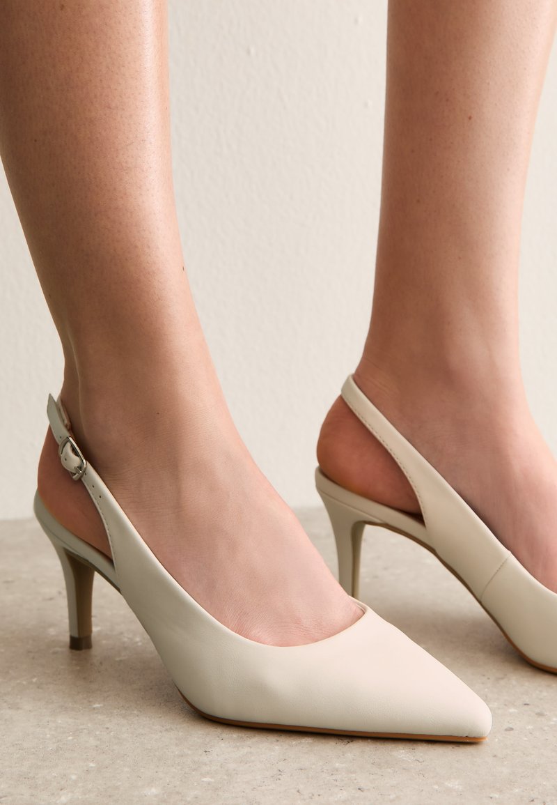 Chaussures à talons kitten beiges avec bride arrière, bouts pointus, portées pieds nus sur un fond clair.