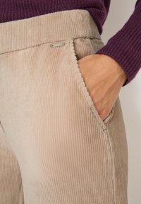 Hand in de zak van beige corduroy broeken, gedragen met een paarse ribgebreide trui.