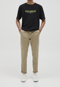 Svart t-shirt med "PULL&BEAR"-logotyp, beige byxor och vita sneakers. T-shirten har lös passform; byxorna är uppvikta vid ärmarna och har dragsko i midjan.