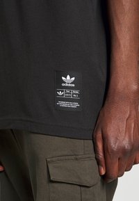Černé bavlněné tričko s obdélníkovou černou značkou s bílým logem Adidas a textem; nošeno s olivově zelenými cargo kalhotami.