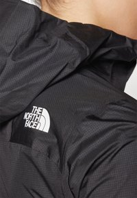 Personne portant une veste noire North Face avec une capuche, montrant le logo sur la zone de l'épaule et une partie du cou visible.