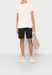 Osoba oblečená v bílém pleteném bezrukávovém svetru přes pruhované tričko, černých kraťasech ke kolenům, béžových teniskách, s béžovou taškou přes rameno.