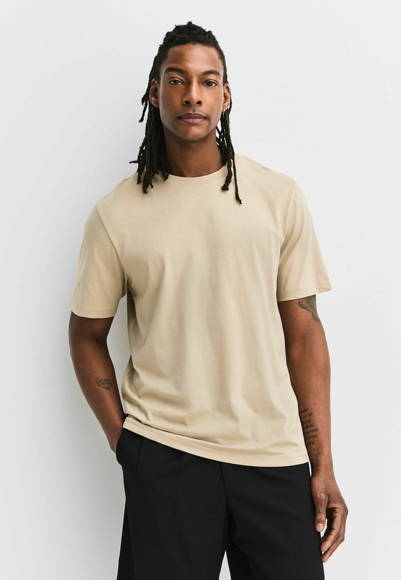 Homme avec dreadlocks portant une chemise beige à manches courtes et un pantalon noir, debout avec une main dans la poche sur un fond clair uni.