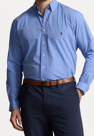 Homme portant une chemise bleu clair à boutons, un pantalon bleu marine foncé et une ceinture en cuir marron usée, une main dans la poche.