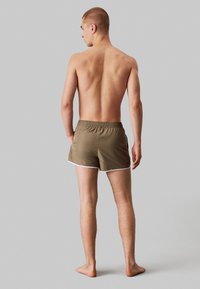 Pantaloni da bagno marroni con bordi bianchi, vita elasticizzata e vestibilità rilassata, visti da dietro su uno sfondo minimalista grigio.