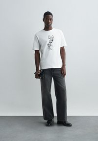 Λευκό βαμβακερό t-shirt με μαύρο καρτούν γραφικό και κείμενο "MAISON KITSUNÉ." Συνδυασμένο με σκούρες γκρι παντελόνες φάρδους και μαύρα παπούτσια.