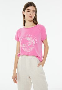 Camiseta de algodón rosa con mangas cortas, cuello redondo y estampado gráfico blanco que presenta un diseño de pájaro y olas. Combinada con pantalones de color claro.