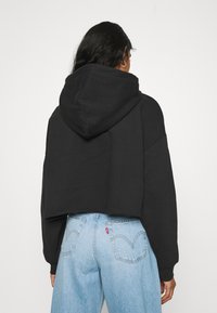 Donna con lunghi capelli neri che indossa un hoodie nero corto e jeans a vita alta di colore blu chiaro, di spalle contro uno sfondo neutro.