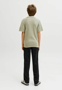 Asmuo su trumpais rudais plaukais, vilkintis šviesiai žalią trumpomis rankovėmis marškinėlį, juodus kelnius ir baltus sportinius batelius, stovintis priešais neutralią foną.