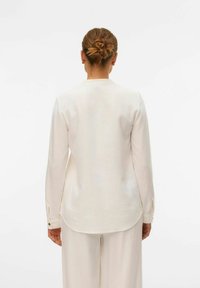 Camicia bianca a maniche lunghe con taglio dritto, polsini abbottonati e un tessuto liscio, leggermente testurizzato. Leggera lucentezza e dettaglio dell'orlo arrotondato.