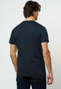 Vīrietis ar tumšiem matiem, valkājot vienkāršu melnu īsroku T-kreklu un tumšas džinsas, redzams no muguras pret vienkāršu gaišu fonu.