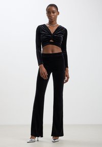Top corto in velluto nero con fronte attorcigliato e maniche lunghe, abbinato a pantaloni a zampa in velluto nero. L'outfit presenta una texture liscia e un design elegante.
