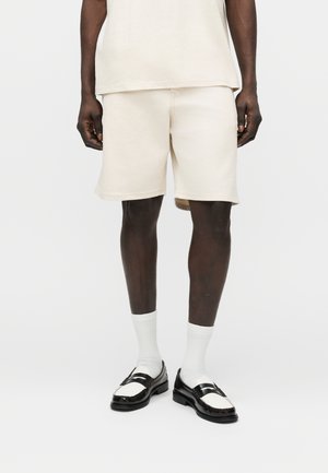 Homme portant un short et une chemise couleur crème, avec des chaussettes blanches et des mocassins noir et blanc, debout devant un fond uni.