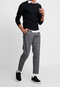Sort striktrøje, hvid skjorte med krave under, grå ternede bukser og sorte sneakers med hvide såler. Outfitet har en afslappet pasform.