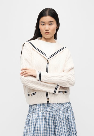 Γυναίκα με μακριά σκούρα μαλλιά, που φοράει κρεμ ζακέτα με πλεκτό σχέδιο και μπλε ρίγες και κουμπιά, καθώς και φούστα με μπλε και λευκό καρό, με τα χέρια σταυρωμένα.