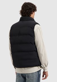 Zwarte puffer vest met een hoge kraag en een gestikte ontwerp, met een gladde textuur. Draagt over een lichtgekleurde, geribbelde shirt.