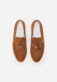 Mocasines de ante marrón con punta redondeada, lengüeta prominente y acentos decorativos de hardware que presentan pequeñas letras metálicas M y H.