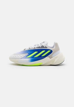 Witte sneaker met blauwe en neongele strepen, dikke zool en witte veters, gezien vanuit zijaanzicht op een effen achtergrond.