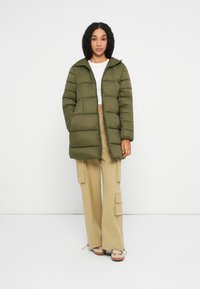 Mujer vestida con una chaqueta acolchada verde oliva, una camiseta blanca de corte corto, pantalones cargo beige y zapatillas beige, de pie contra un fondo blanco liso.