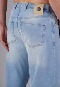 Hellblaue Jeans aus Denim mit einer verblichenen Textur, ausgestattet mit einem Etikett am Bund und zwei Gesäßtaschen mit kupferfarbenen Nieten.