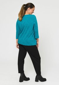 Zeleno modra majica z teksturirano valovito vzorcem, s trečjino rokavi, v kombinaciji z črnimi hlačami in gležnjarji, prikazana s hrbtne strani.